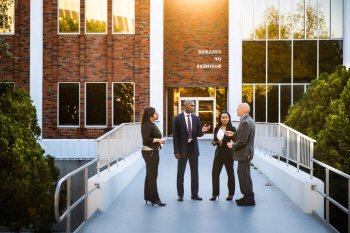 Four people standing outside of a brick building and conversing. The building features a sign that reads "College of Business."